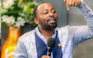A medium shot of Bishop Ben Owusu speaking into a microphone while gesturing emphatically with his hand. He is dressed in a patterned light blue and navy shirt, with a blurred background featuring floral decorations.