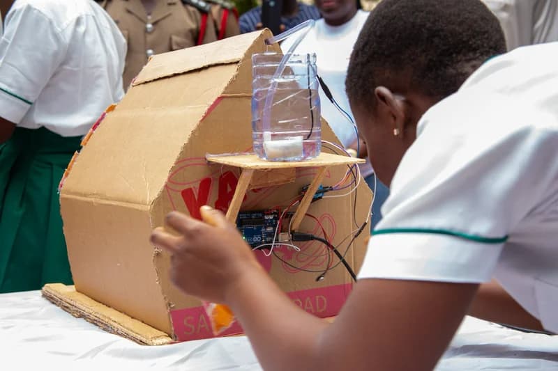 A student in a white and green uniform works on a STEM project featuring a cardboard house model integrated with an Arduino microcontroller and a water pump system. The hands-on demonstration appears to be part of a technical workshop or exhibition, with other students and a uniformed official visible in the background.