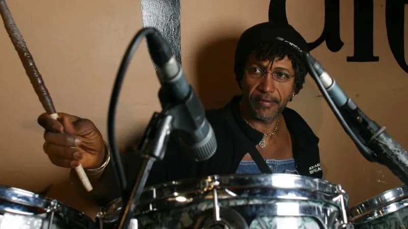 A close-up photograph of legendary reggae drummer Sly Dunbar seated at a drum kit with microphones positioned in front of him. He is wearing a black beanie, glasses, and a denim outfit while holding a drumstick in a studio-like environment.