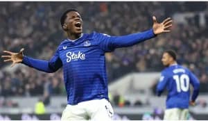 Everton player Louie Barry celebrates with arms outstretched and an ecstatic expression after scoring a goal during a match. He is wearing the club's blue home jersey with the Stake sponsor logo, while a teammate and a blurred crowd of spectators are visible in the background.