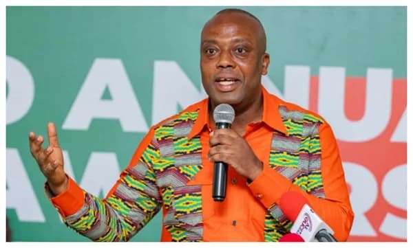 Dr. Mohammed Amin Adam, Ghana's Minister for Finance, is captured speaking into a microphone during a public address. He is wearing a vibrant orange shirt featuring intricate Kente-styled patterns on the sleeves and front panels, gesturing with one hand as he speaks.