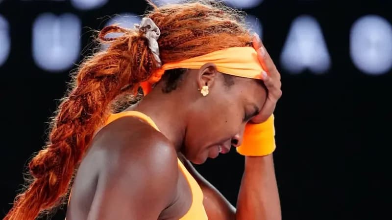 A close-up side profile of professional tennis player Coco Gauff looking visibly distressed and frustrated on the court. She is wearing a bright orange headband and matching top, with her hand pressed against her forehead while her hair is styled in long, reddish-brown braids.