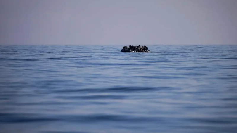 An overcrowded inflatable boat carrying dozens of individuals is seen adrift in the middle of a vast, calm blue ocean. The horizon is clear under a pale sky, emphasizing the isolation and precarious nature of the vessel's journey.