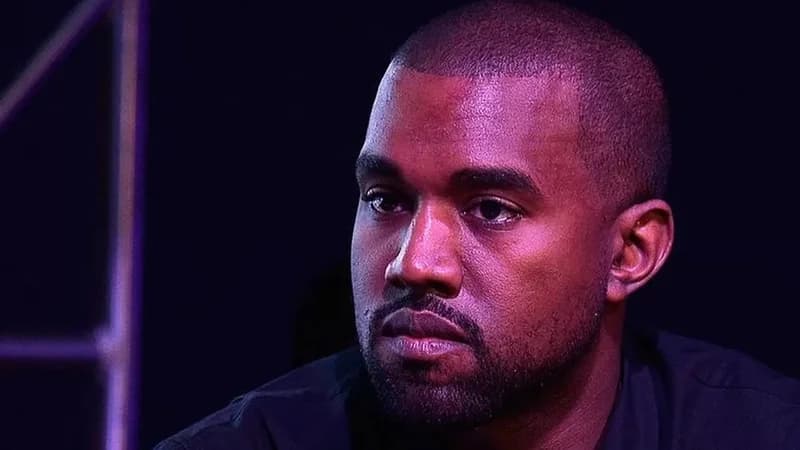 A close-up portrait of Kanye West, also known as Ye, appearing with a serious and pensive expression. The image features dramatic purple and pink lighting cast over his face against a dark, out-of-focus background.
