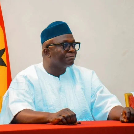 A portrait photograph of Bryan Acheampong, a Ghanaian politician and Member of Parliament. He is captured seated at a table with a red cloth, wearing a traditional white smock, a teal-colored cap, and black-rimmed glasses, with a portion of the Ghana flag visible in the background.