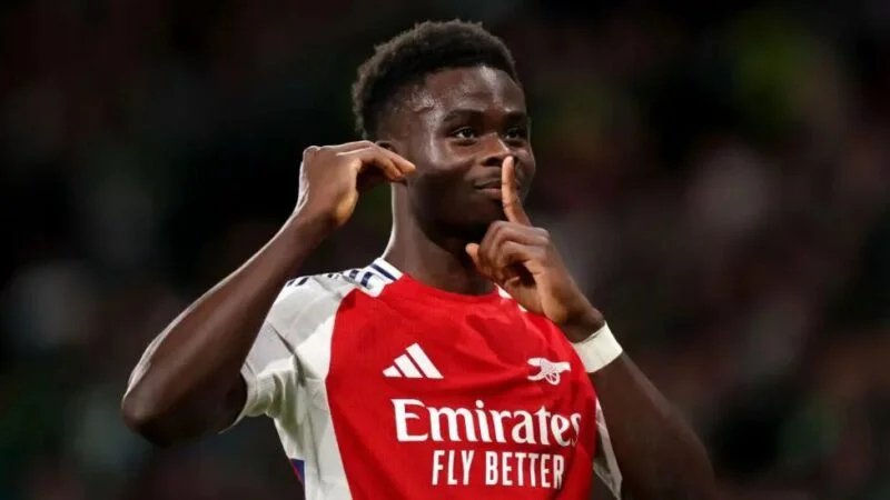 Arsenal footballer Bukayo Saka is shown celebrating in the team's red and white home jersey. He is making a shushing gesture with one finger over his lips while holding his other hand to his ear against a blurred stadium background.