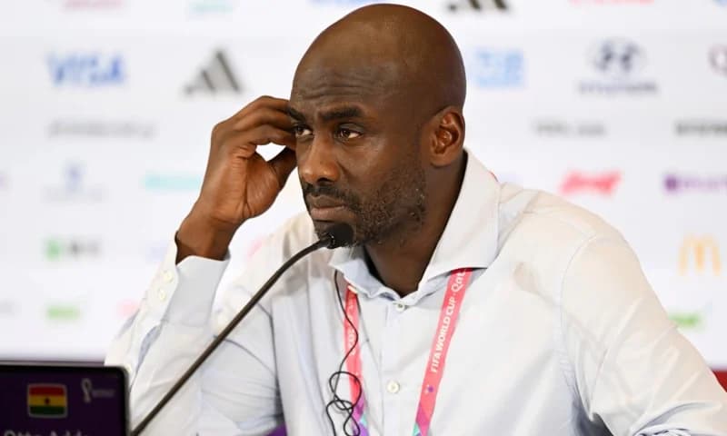 Otto Addo, the head coach of the Ghana Black Stars, is pictured at a press conference during the FIFA World Cup. He appears pensive, resting his hand near his ear while seated behind a microphone and a backdrop featuring official tournament sponsors.