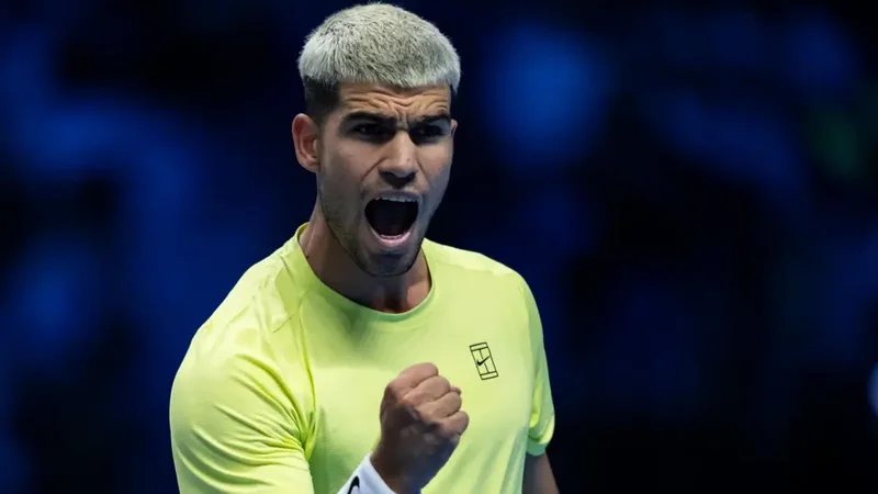 Spanish tennis star Carlos Alcaraz is captured in a moment of intense celebration during a match. He is seen shouting with a clenched fist, wearing a bright neon yellow jersey and sporting a bleached hairstyle against a dark blue background.