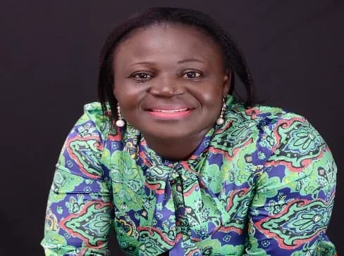 A portrait photograph of Princess Osisiadan-Quaye, who is recognized for her work in Ghana's health insurance sector. She is smiling at the camera, wearing a colorful green and blue patterned blouse with a pussy-bow neckline against a solid black background.