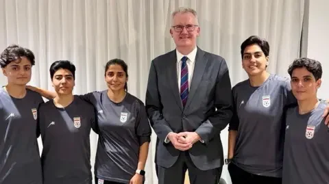 Reuters Australian Home Affairs Minister Tony Burke, wearing a grey suit, poses with the five Iranian football players who have been granted humanitarian visas. They are all smiling and wearing grey football jerseys.