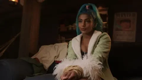 Netflix Chandran in character as Nefertari Vivi wears a green coat with fluffy sleeves and has blue hair tied up in a ponytail with a metallic headband