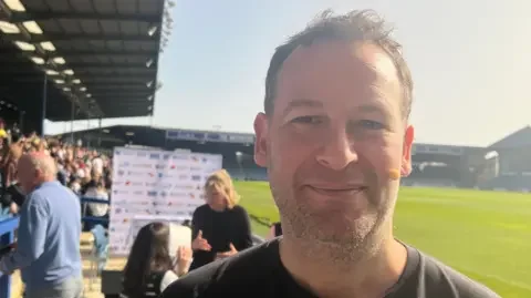 Dan Freedman looking at the camera with a football pitch and crowds in the background. Dan is wearing a microphone on his face.