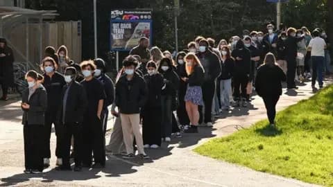 Reuters People queue to receive vaccinations at the sports centre at the University of Kent campus in Canterbury. They are all wearing masks and are lined up in the sunshine near an area of grass. There are trees behind them and signposts and there is a student noticeboard with a poster.