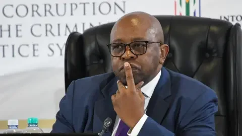 Gallo Images via Getty Images Mbuyiseli Madlanga in glasses, a dark blue suit and purple tie sits on a black chair as he listens during a hearing of the Madlanga Commission with one of his forefingers pressed to his lips.