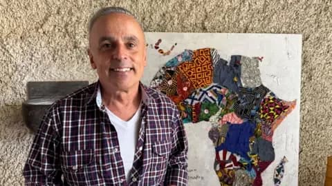A Spanish man in his later middle age wears a checked shirt. He smiles in front of a map of Africa made out of fabric.