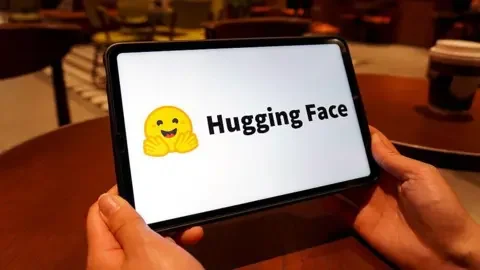 Getty Images The Hugging Face logo shown on a tablet screen. It is a yellow emoji of a smiling face with its hands spread out in front of it.