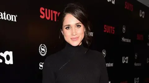 Getty Images The Duchess of Sussex Meghan Markle stands in front of a promotional board with Suits written on it in red writing. She is wearing a black dress and has her dark hair tied up.