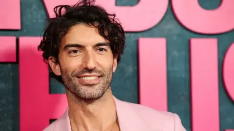 Reuters Justin Baldoni in a pink suit posing in front of a grey backdrop with large pink letters at a film premiere