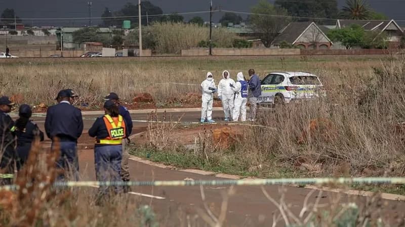 A crime scene investigation in an open field in South Africa, featuring forensic experts in white hazmat suits and police officers. A marked police vehicle is parked in the background while investigators gather evidence behind yellow cordoned-off tape.