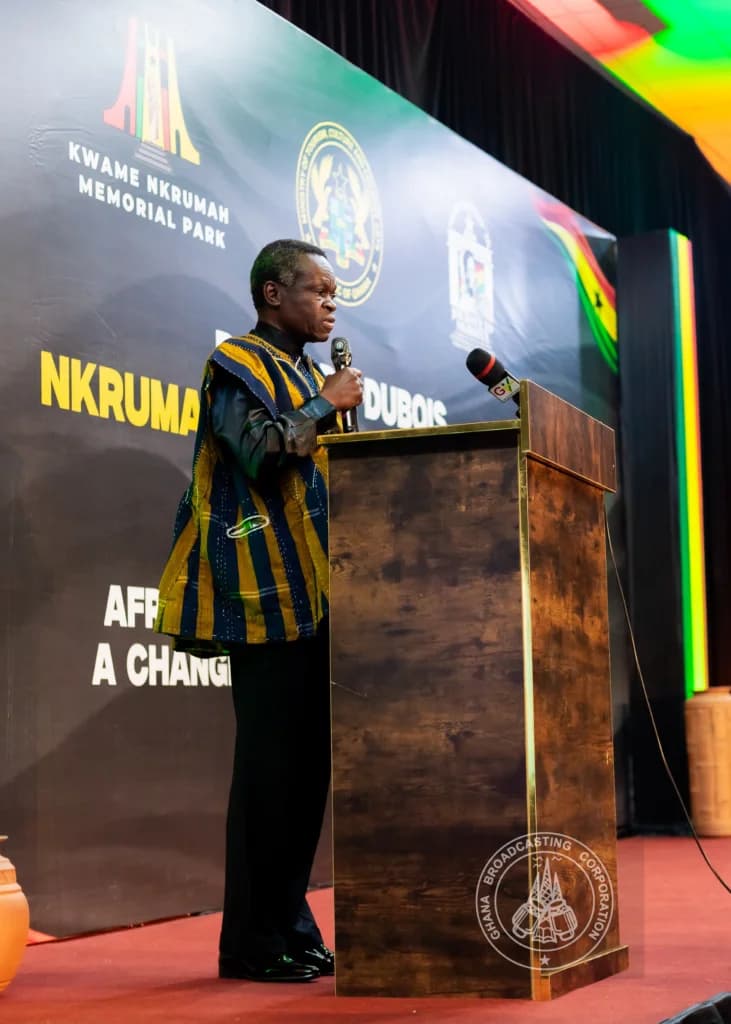 Professor P.L.O. Lumumba is pictured delivering a speech from a wooden lectern at an event in Accra, Ghana. He is dressed in a traditional blue and gold striped smock, standing before a backdrop that features the Kwame Nkrumah Memorial Park logo and official government emblems.
