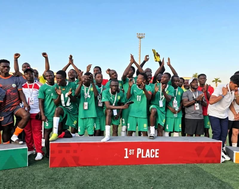 The University for Development Studies (UDS) football team celebrates their victory on a winners' podium. The players are wearing green jerseys and white socks, cheering and clapping behind a red banner marked '1st PLACE' on a football field under a clear blue sky.