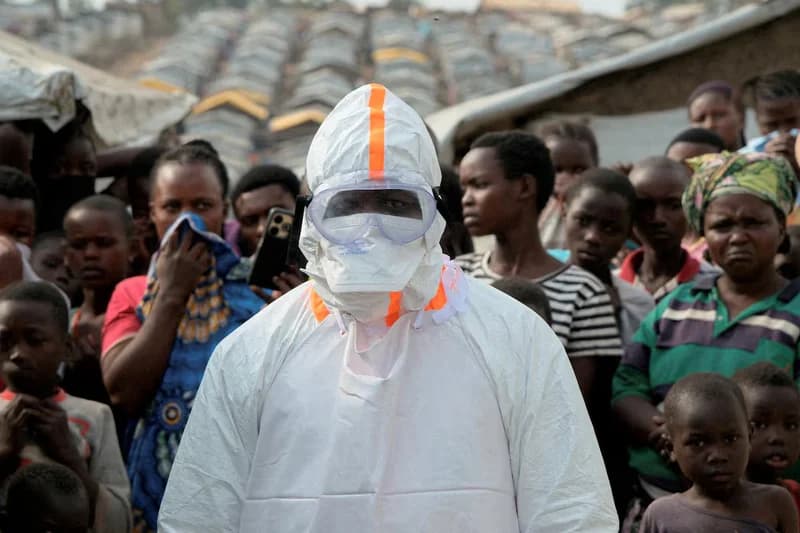 A health worker dressed in full white personal protective equipment (PPE) with orange accents, including a face mask and goggles, stands in the foreground of a crowded outdoor area. A large group of people, primarily children and young adults, are visible behind the worker, set against a backdrop of numerous temporary shelters in what appears to be a camp or informal settlement.