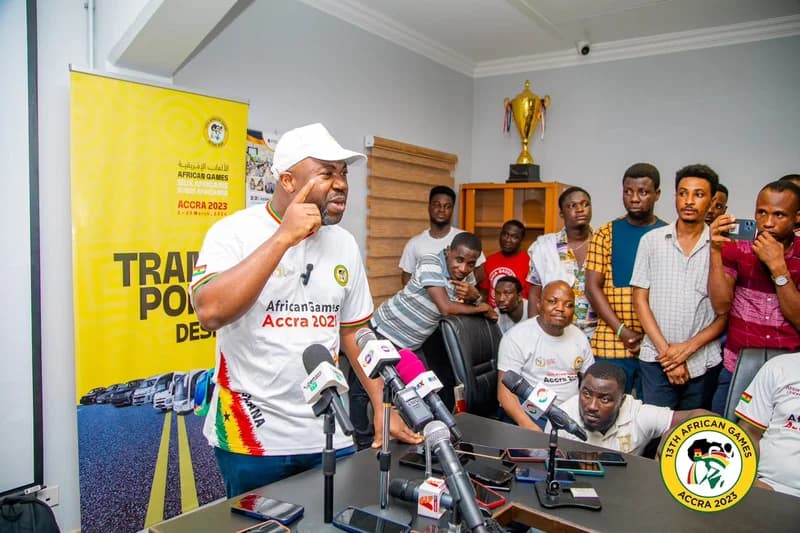 Samson Deen, President of the African Paralympic Committee, is seen speaking at a press conference regarding the 13th African Games held in Accra. He wears branded apparel for the games and gestures emphatically while standing behind a table crowded with news microphones, surrounded by observers.