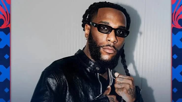 A portrait of Nigerian Afrobeats star Burna Boy wearing a black leather jacket and dark sunglasses. He is positioned against a plain light gray background, with decorative blue and red geometric borders on the sides.