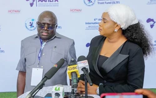 Nana Kwaku Agyei Yeboah, CEO of the Students Loan Trust Fund (SLTF), speaks to the media at the 6th AAHEFA International Conference. He is flanked by a female colleague while several news microphones from outlets like GBC are positioned in front of them to capture his statement.