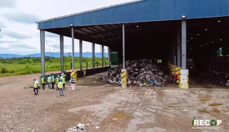 The image shows a large industrial shed housing a massive pile of waste, with a group of about a dozen people in high-visibility vests standing on the dirt ground outside. The facility is situated in a rural area with green vegetation and hills visible in the background, illustrating the scale of waste management infrastructure in the region.