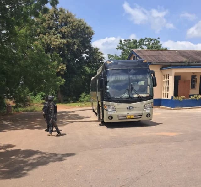 A tan Kia bus is parked in front of a building with blue trim, while two armed security personnel in camouflage uniforms walk across the foreground. The scene likely depicts the arrival or transport of suspects at a legal or police facility in Ghana.