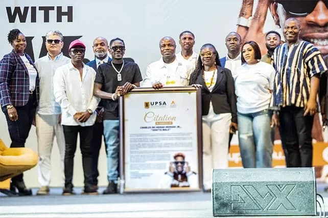 Ghanaian dancehall musician Shatta Wale stands with university officials on a stage, holding a large framed citation presented to him. The group is posing for a commemorative photograph during an entrepreneurship conference at the University of Professional Studies, Accra (UPSA).
