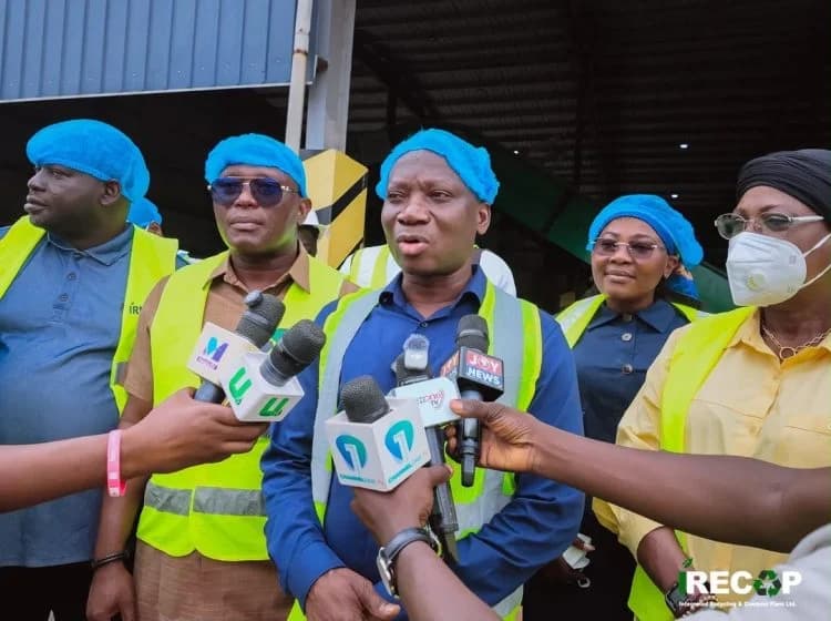 A group of officials wearing blue hairnets and yellow safety vests address members of the press during a site visit. Several microphones from news outlets like Joy News and Channel One TV are positioned in front of the primary speaker at what appears to be a waste management facility.