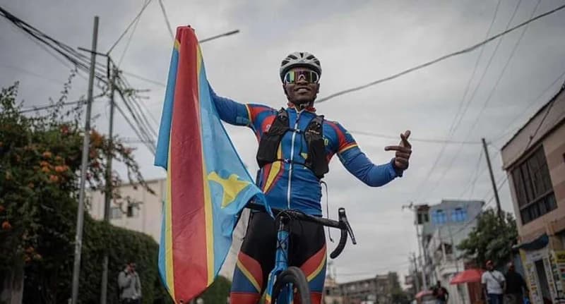 A Congolese cyclist, identified as Gedeon Ngwendema, smiles while holding the flag of the Democratic Republic of the Congo and standing with his bicycle. He is wearing professional cycling gear including a helmet, sunglasses, and a jersey featuring the national colors against an urban street background.