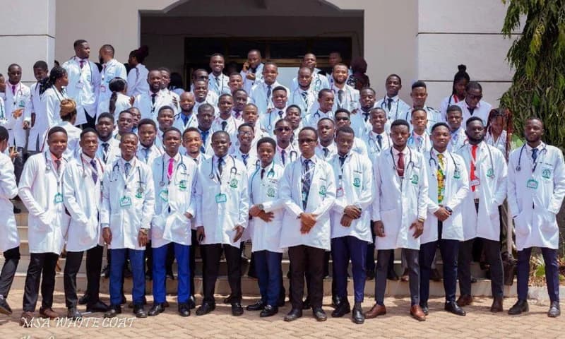 A large group of Ghanaian medical graduates posing for a formal group photograph in front of a building. They are all dressed in white medical lab coats and formal attire, with many wearing stethoscopes, representing a cohort of newly trained healthcare professionals.