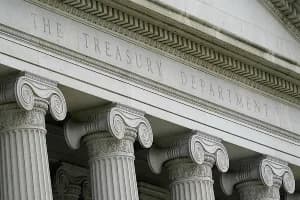 A close-up photograph of the classical stone facade of the United States Treasury Department building in Washington, D.C. The image highlights the detailed Ionic columns and the institution's name engraved prominently in the architecture.