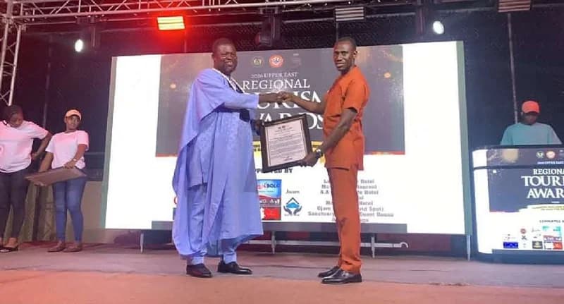 The image shows a man in a traditional light blue smock, presumably Michael Adangba, receiving a framed award from an official in an orange outfit. They are standing on a stage in front of a large digital backdrop for the 2026 Upper East Regional Tourism Awards.