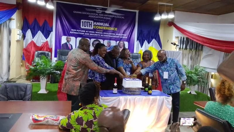 A group of officials and attendees participate in a cake-cutting ceremony during the 10th Annual Conference of the Ghana Sociological and Anthropological Association (GSAA). The event features a large purple banner with the conference theme focusing on social exclusion and resource extraction in Africa.