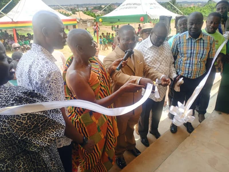 A group of officials and traditional leaders are seen during a ribbon-cutting ceremony for the commissioning of new school blocks. A man in a suit cuts the ceremonial white ribbon with scissors while holding a microphone, flanked by a traditional chief in vibrant kente cloth. In the background, school children and community members gather under event tents for the occasion.
