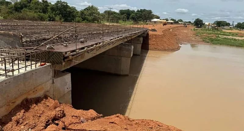 A bridge currently under construction spans a wide, murky river with high water levels. The structure features concrete supports and a deck reinforced with exposed metal rebar, surrounded by red earth and rural vegetation.