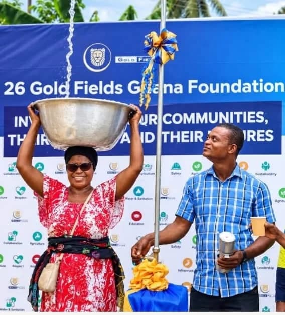A woman balances a large silver basin on her head as water flows into it from a newly commissioned standpipe. A man in a blue checkered shirt stands beside her, observing the water flow in front of a Gold Fields Ghana Foundation banner.
