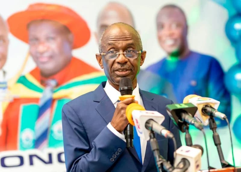 Johnson Asiedu Nketiah, the National Chairman of the National Democratic Congress (NDC), is shown speaking into multiple microphones at a press event. He is wearing a dark blue suit and spectacles, appearing to deliver a serious address against a blurred background featuring other figures and colorful elements.