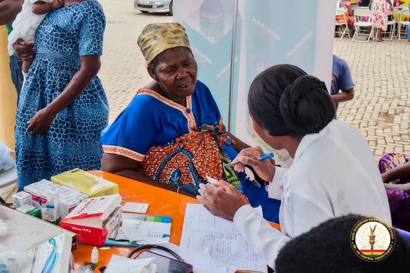 A healthcare professional in a white lab coat is seen consulting with an elderly woman during a community health outreach event. The scene features a table stocked with medical supplies, including rapid diagnostic test kits, signifying an active health screening session in Ghana.