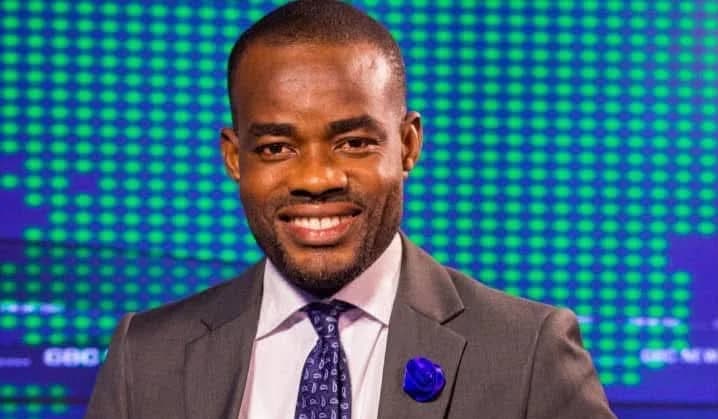 A headshot of GBC sports journalist Theophilus Sampah smiling while wearing a grey suit, white shirt, and a blue patterned tie with a blue flower lapel pin. He is standing in front of a blue and green digital background typical of a television news studio.