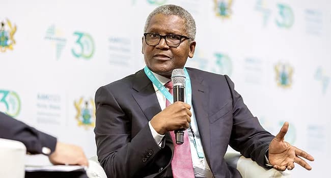 Nigerian industrialist Aliko Dangote is captured speaking into a microphone during a panel discussion. He is dressed in a formal dark suit and red tie, gesturing with his hand against a backdrop featuring Afreximbank 30th-anniversary branding and the Ghanaian coat of arms.