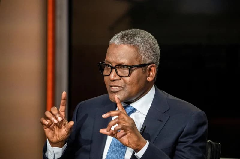 A professional portrait of Aliko Dangote, the prominent Nigerian billionaire and businessman, gesturing with his hands during an interview. He is seen wearing a navy blue suit, a white shirt, a patterned blue tie, and black-framed eyeglasses against a dark, studio-like background.