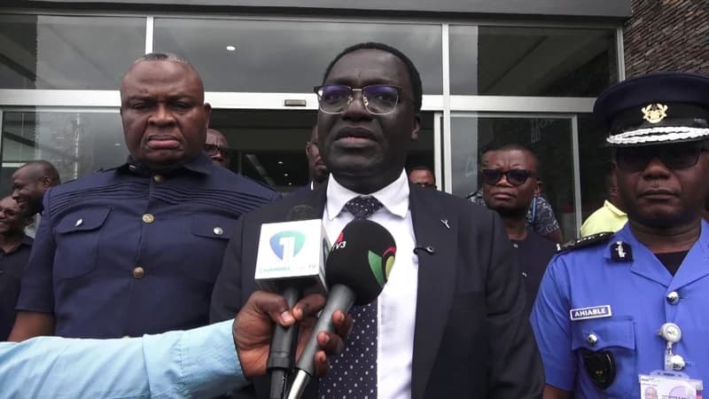 A man in a formal suit and glasses addresses the press, flanked by officials including a uniformed officer from the Ghana Revenue Authority's Customs division. The scene captures a media briefing with microphones from TV3 and Channel One TV visible in the foreground.