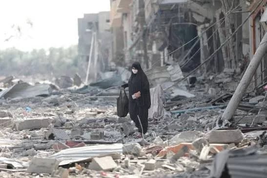 A woman wearing a black abaya and a white face mask walks through a street heavily laden with rubble and the ruins of destroyed buildings. The image captures the devastation of a conflict zone, highlighting the struggle of civilians navigating a landscape of war.