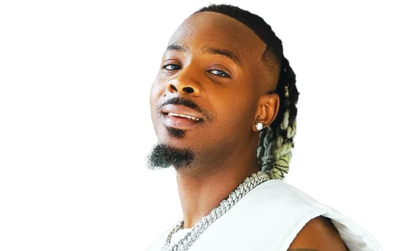 A professional portrait of Nigerian singer and producer Young Jonn, featured against a plain white background. He is shown from the chest up, wearing a white sleeveless top and a prominent silver cuban link chain.