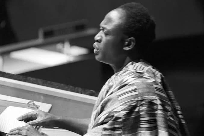 A black and white historical photograph showing Kwame Nkrumah, Ghana's first president, speaking from a podium. He is captured in a side profile, wearing a traditional Kente cloth while delivering a speech at what appears to be an international assembly.